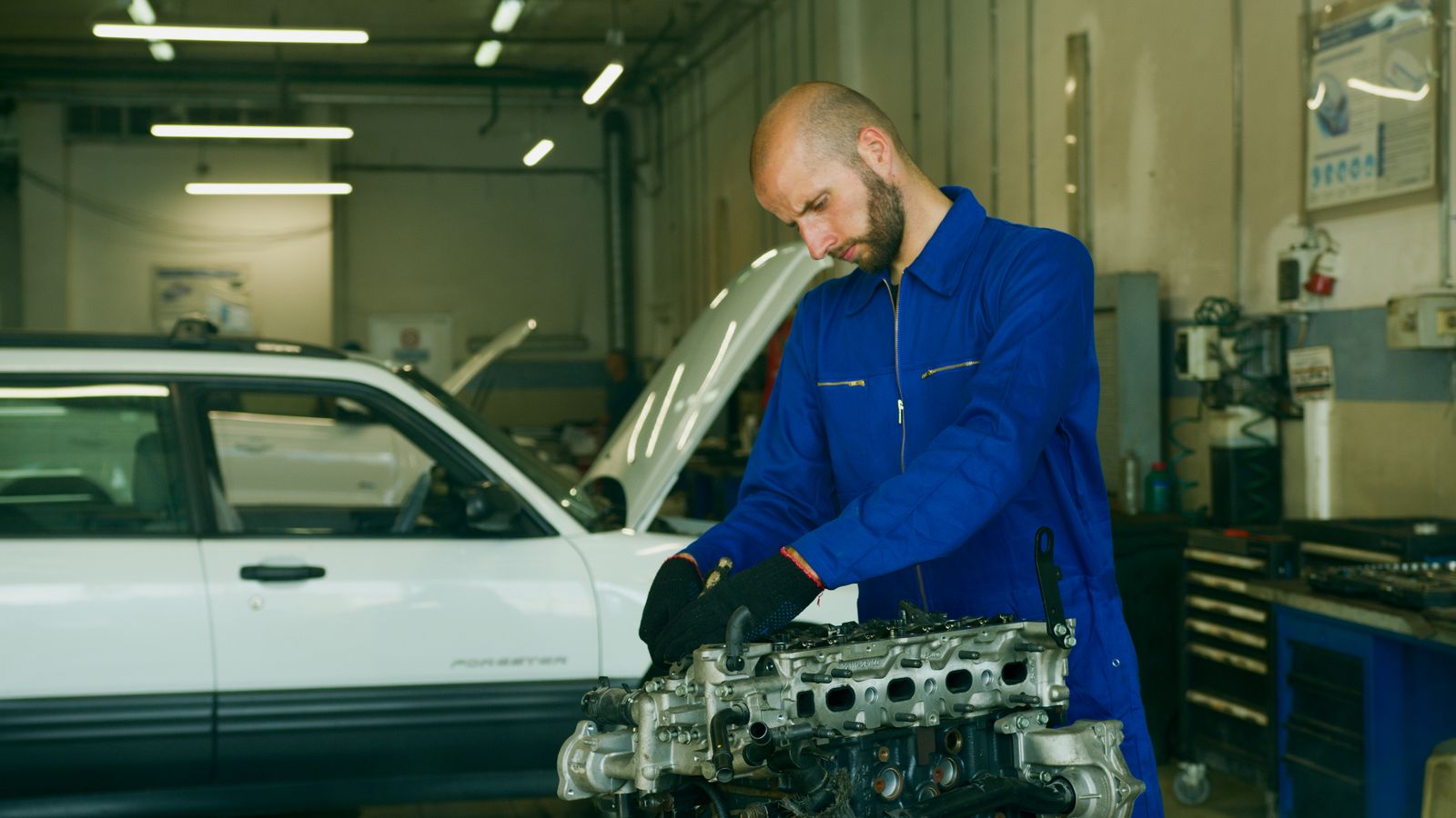 Mecânico em um macacão azul inspecionando motor de carro em uma oficina de garagem.